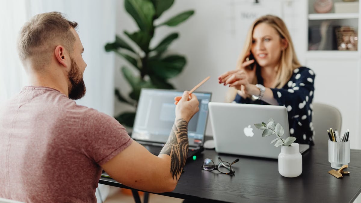 Man and woman discussing work at a table with laptops in a modern office.